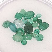 Top view of a collection of twenty loose emerald gemstones in various sizes and shapes on a white background.