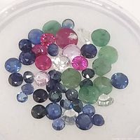 Top view of loose mixed green, red, blue, clear, and pink faceted round and oval gemstones in a shallow container.