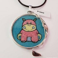 Front of pendant showing pink hippopotamus design with blue background in silver frame on a black cord.