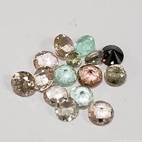 Photo of 14 loose faceted gemstones in various colors including pink, green, brown and a black stone scattered on white background for size and detail reference.