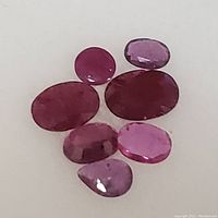 Seven loose faceted ruby gemstones in varying shades of red and purple, various cuts including oval, round, and pear shapes.