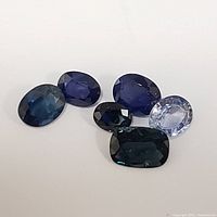 Six loose sapphire gemstones of different shapes and shades arranged on a white background.