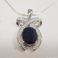 Front view of a silver pendant necklace featuring a large oval blue sapphire-like stone surrounded by small cubic zirconia stones set in a ribbon shaped design.