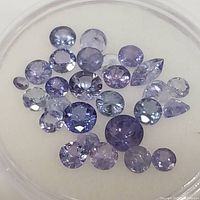 Overhead view of 25 loose tanzanite gemstones in round and pear cuts with various violet-blue shades in a clear container.
