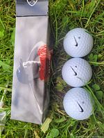 Three white Nike golf balls numbered 2, 4, and 2 with black swoosh logos next to a black box featuring an image of Tiger Woods