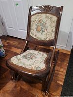 Front view of antique wooden folding rocking chair with floral tapestry seat and backrest, wood carved details visible.