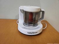 White Baby Brezza baby food maker showing base with buttons and transparent blending container with handle and red blade inside.