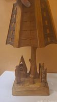 Front view of the carved wooden lamp showing the woman, small house/oven, tree trunk lamp post, and wooden shade with square cutouts.