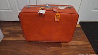 Orange hard-sided rectangular suitcase with metal lock and handle shown closed on wooden floor.