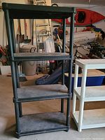 Full view of black plastic four-tier shelf unit showing all shelves and cylindrical legs. Visible dust and dirt on shelves.