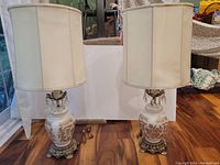 Full view of both vintage lamps on hardwood floor against white background showing white fabric shades and ornate bases.
