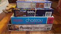Stack of 4 jigsaw puzzle boxes showing titles and brands: Puzzle's Collection, Chateau, 500-piece puzzle, and another Puzzle's Collection.