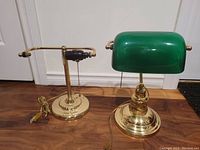 Two vintage bankers desk lamps side by side. One lamp has a green glass shade, the other has no shade. Both lamps have brass bases and pull chains.