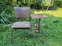 Full view of vintage chair with tubular metal frame, beige textured fabric seat and back, attached small side table with wood-grain top, and wire magazine holder beneath the tabletop.