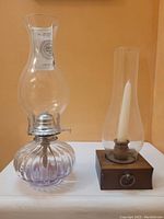 Both items together on display with orange background. Left is ribbed glass oil lamp with chimney and attached instructions. Right is wooden candle holder with glass chimney and white taper candle.