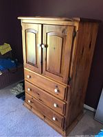 Wood cabinet armoire in a natural finish showing upper double panel doors and three drawers below on carpeted floor.