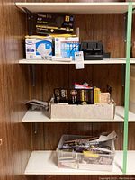 Three shelves showing multiple boxes of light bulbs, a black battery charger, plastic container with batteries and office supplies.
