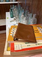 Photo showing a collection of vintage glass bottles in various sizes and colors, alongside vintage printed catalogs and a brown binder.