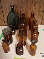 Group photo of 12 vintage glass bottles including amber bottles and one dark green bottle with lids on some bottles.