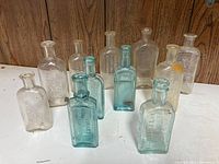 Photo showing 11 vintage glass bottles arranged on a white surface against a wood panel background with clear and aqua blue colors, some with embossed text.