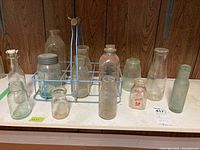 Full view of 15 pieces: a blue metal bottle carrier holding several vintage glass bottles, plus additional bottles arranged around it on a white surface against wood panel background.