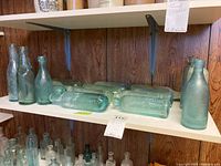 Multiple aqua glass bottles on white shelving against wood panel background