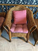 Full view of the wicker chair showing the woven wicker design, wooden armrests and legs, with pink cushions on seat and back.