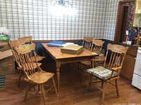 Six wooden pressback chairs around a wooden dining table in a dining room with plaid wallpaper. Two of the chairs have armrests, four do not. The chairs have a natural wood finish.