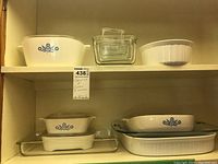 Photo of two shelves with various CorningWare casseroles and glass bakeware including some with lids and a white round fluted baking dish