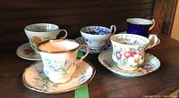 Shows 6 cups and saucers with various floral designs and colors on a wooden shelf.
