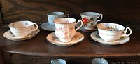 Five assorted vintage cups and saucers arranged on a wooden surface showing varied floral and scenic designs with gold accents.
