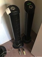 Two black Seville Classics tower fans standing upright against a wall, each with a remote control placed in front on the floor.