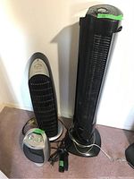 Two black tower fans standing upright next to each other with cords and remotes. Taller fan on right with control panel on top.