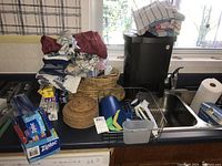 Countertop with stacked kitchen towels and aprons, assorted placemats and trivets, dish drainer with cleaning tools, and paper goods such as Ziploc bags.