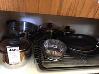 Various stainless steel pots with glass lids and black handles, a perforated metal steamer insert, dark metal baking pans stacked, and a chrome wire rack inside a wooden cabinet.