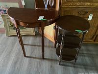 Two wooden side tables shown together on floor: one demi-lune with drawer, one round three-tiered.