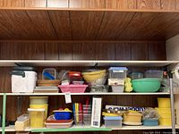 Photo showing two shelves with a variety of Tupperware containers, plastic storage boxes, mixing bowls in green and yellow, springform pans, woven baskets and other kitchen accessories.