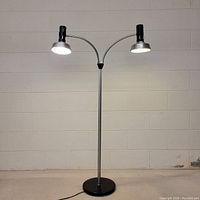 Full view of the lamp showing black circular base, metal central pole, and two black lamps on gooseneck arms.