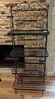 Front view of black metal bakers rack with ornate scroll design against stone wall.