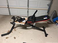 Full view of the workout bench with black and red padding, metal frame, a pair of gloves placed on a wooden platform, and some additional parts.