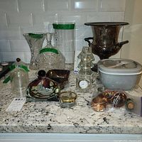 Front view of the entire lot on a granite countertop showing all glassware, clock, silverstone carafe, candle holders, napkin rings, photo frame, and glass bell domes.