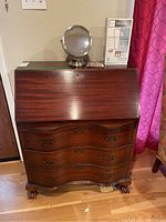 Secretary desk closed showing polished wood finish, drop-down front panel, three curved drawers with metal handles, and ball and claw carved feet. Vanity mirror and boxed task lamp on top.