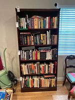 Front view of the dark wooden bookcase filled with books, placed near a window and some furniture.