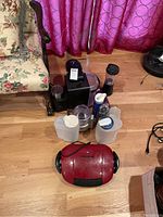 Red George Foreman grill with black handles and plastic jugs and coffee maker on wood floor.