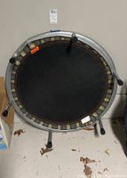 Top view of round mini trampoline showing black fabric surface, elastic bands with metal frame and support legs, some dirt and pet hair on floor under it.