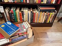 Photo showing a shelf filled with assorted paperback and hardback books behind a box filled with more books, including travel guides and popular fiction titles.