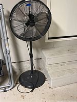 Full view of black pedestal fan showing base, pole, fan head with three silver blades enclosed in black wire cage, placed indoors near stairs.