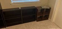 Two dark wood bookshelves positioned side by side in front of a window and wall, with one visibly larger than the other, both showing rectangular open shelving.