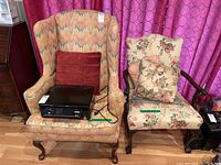 Two side chairs shown side-by-side: a flame stitch wing back chair with red pillow and a floral accent chair with carved wooden arms and floral pillow placed in front of purple patterned curtains on hardwood floor.