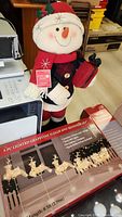 Close-up of plush snowman figure holding a red gift and the boxed 5-piece lighted grapevine sleigh and reindeer set
